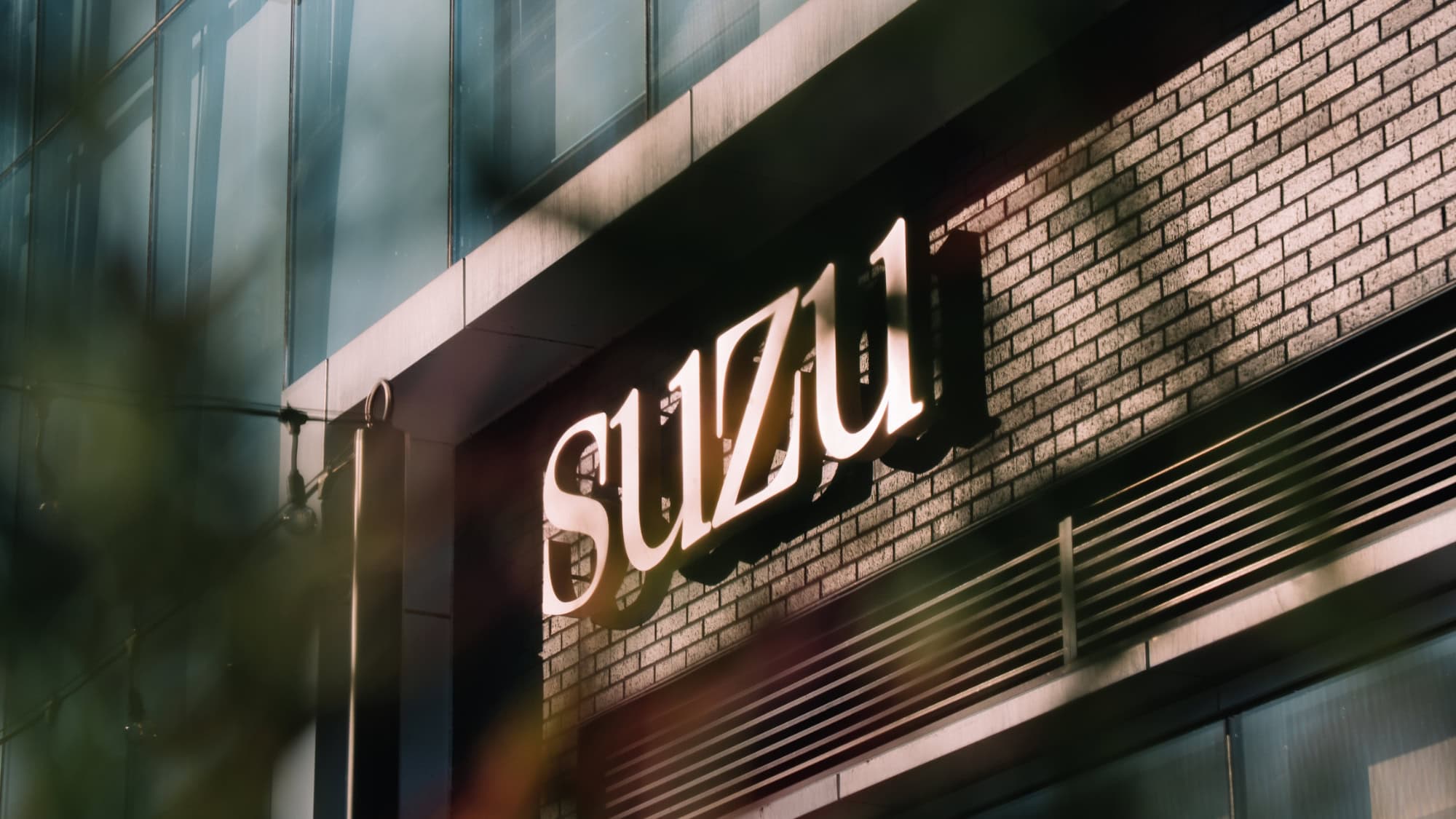 Illuminated 3D sign reading “SUZU” mounted on a brick exterior wall.
