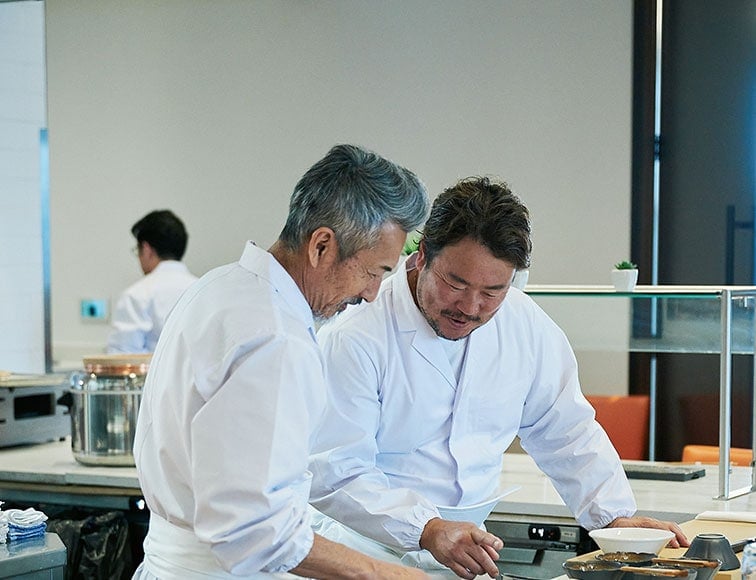 Two chefs in white uniforms work together in a modern kitchen, focusing on food preparation at the counter.