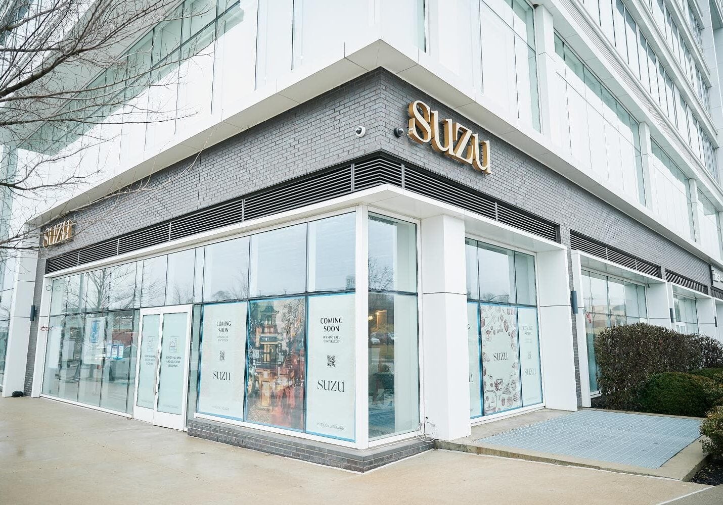 Corner view of a modern building with large windows, SUZU signage, and posters on the glass announcing the restaurant’s upcoming opening.
