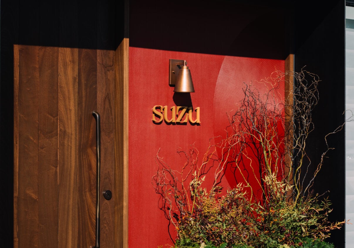 The word “SUZU” in gold lettering on a red textured wall, next to a wooden door and a metal light fixture.
