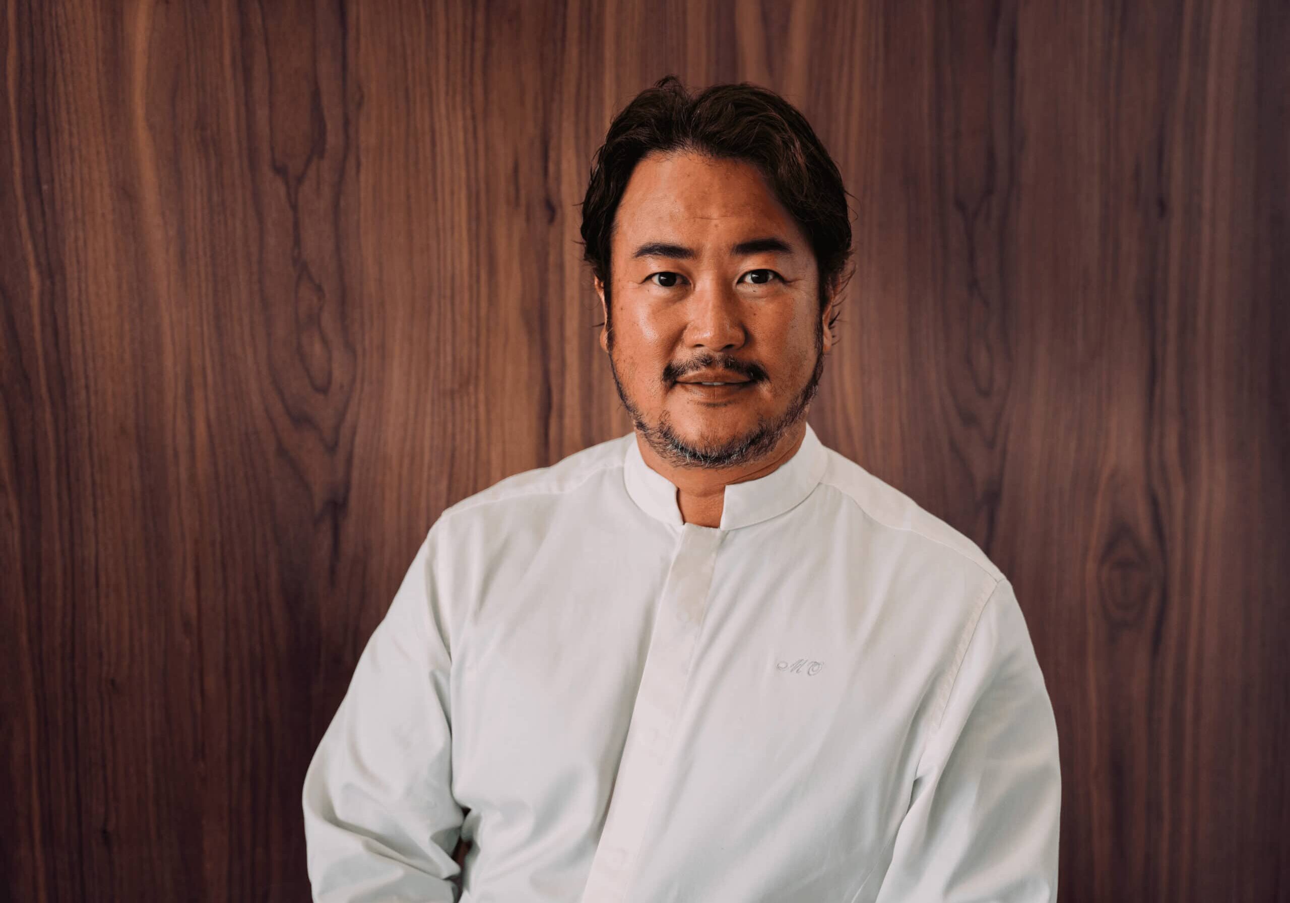 A man with short dark hair and a trimmed beard wears a white button-up shirt, standing in front of a wooden wall and looking at the camera.
